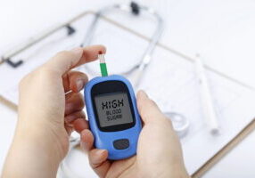 Hand holding a blood glucose meter measuring blood sugar, the background is a stethoscope and chart file